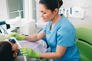 A pediatric dentist wearing green gloves adjusts an inhalation sedation mask over a young boy's nose during a dental procedure.