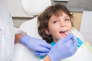 A young boy smiling happily while lying in a pediatric dental chair during a dental examination as a dentist uses a mouth mirror.