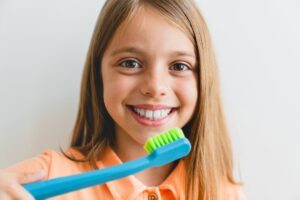A smiling young girl with long brown hair holding up a bright blue and green toothbrush to her teeth.