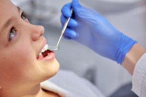 A close-up side profile of a young girl smiling while a pediatric dentist wearing blue gloves gently uses a dental mirror to examine her teeth.