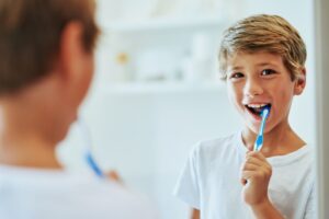 A cheerful young boy smiling at his reflection in a bathroom mirror while brushing his teeth with a blue toothbrush.