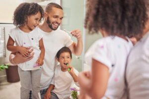 A father laughing and holding his young daughter while his son brushes his teeth in front of a bathroom mirror, showing a fun way to prepare a child for a dental visit.