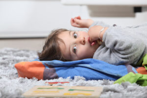 A young girl lying on a grey shag rug and sucking her thumb, showcasing a common childhood habit that can impact dental development.