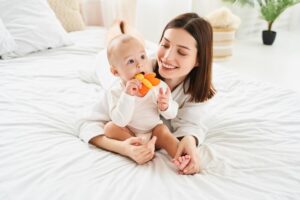 A smiling mother lying on a white bed while holding her baby boy, who is happily chewing on an orange butterfly-shaped teething toy.