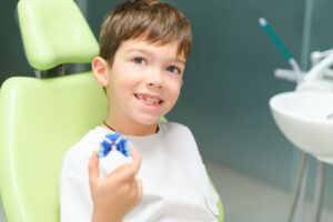 A smiling young boy sitting in a green pediatric dental chair while holding up a blue orthodontic removable appliance (retainer).