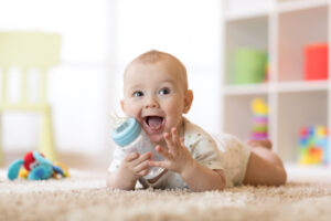 A happy, smiling baby boy lying on a carpet and holding a baby bottle.