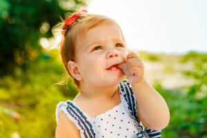 A happy, smiling toddler girl with a red bow in her hair sitting outdoors and sucking her thumb.
