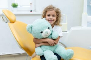 A smiling young girl sitting in a pediatric dental chair and happily hugging a large, soft teal teddy bear during her visit.