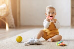 A happy, blonde toddler sitting on a woven rug and playing with colorful teething toys.