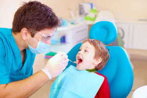A smiling male pediatric dentist wearing safety glasses and a surgical mask gently examines a young boy's teeth during a dental checkup.