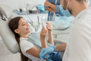 A young girl sitting in a dental chair smiles happily as she looks at her teeth in a handheld mirror held up by a pediatric dentist.
