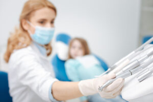 A pediatric dentist in a surgical mask and gloves handles a dental drill while a young girl sits in the background blurred in the dental chair.